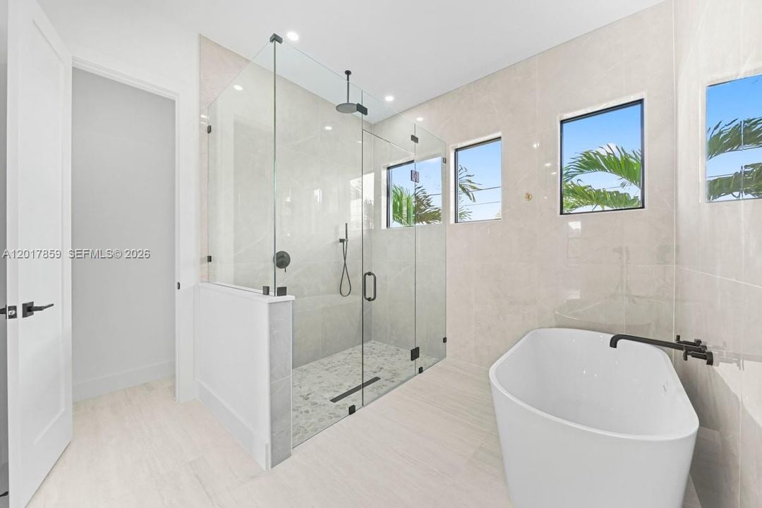 Walk-in glass-enclosed shower with rain-style showerhead and handheld attachment, freestanding soaking tub with matte black wall-mounted faucet, floor-to-ceiling tile wall finishes, and light-toned flooring