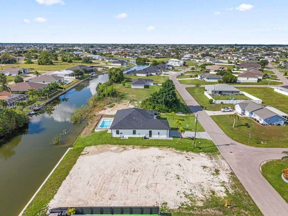 Aerial perspective of a single-story residence featuring a private canal-side swimming pool, a concrete driveway, and extensive undeveloped lot space