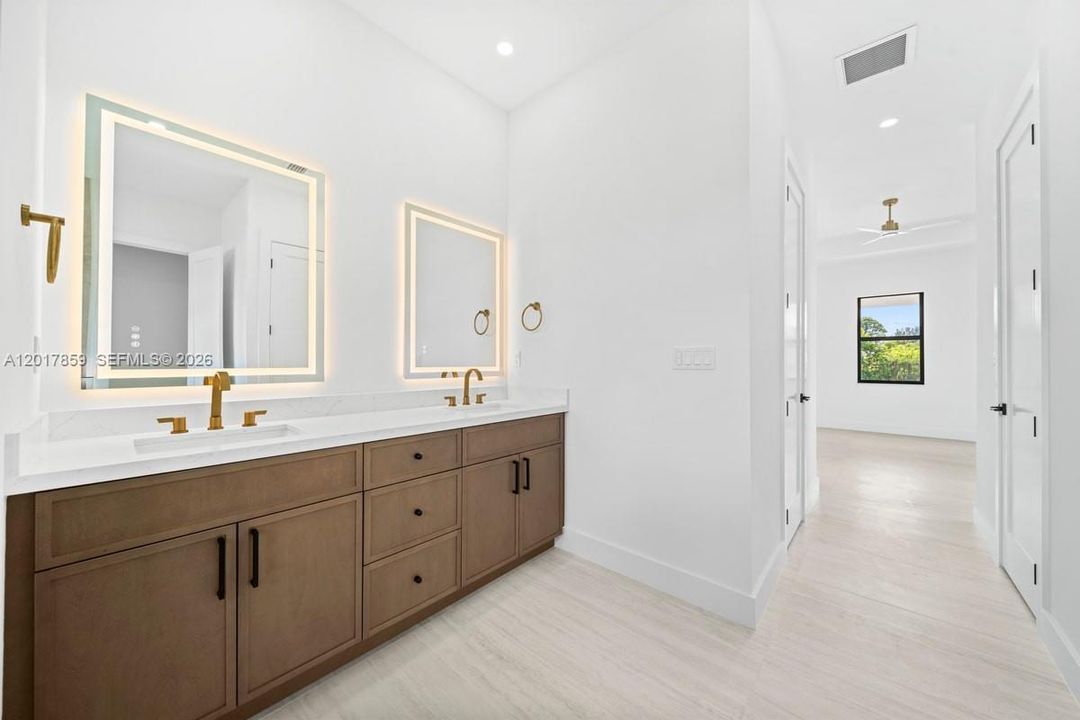 Double vanity featuring wood-finish cabinetry, white solid-surface countertop, integrated rectangular sinks, brass-toned hardware, and rectangular backlit mirrors