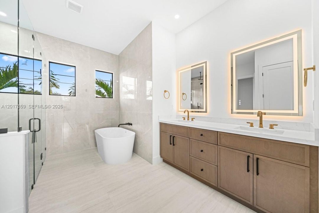 Dual-sink vanity with wood-finish cabinetry, quartz countertops, and brass-tone fixtures, complemented by a freestanding soaking tub, floor-to-ceiling tile work, and an expansive walk-in glass-enclosed shower