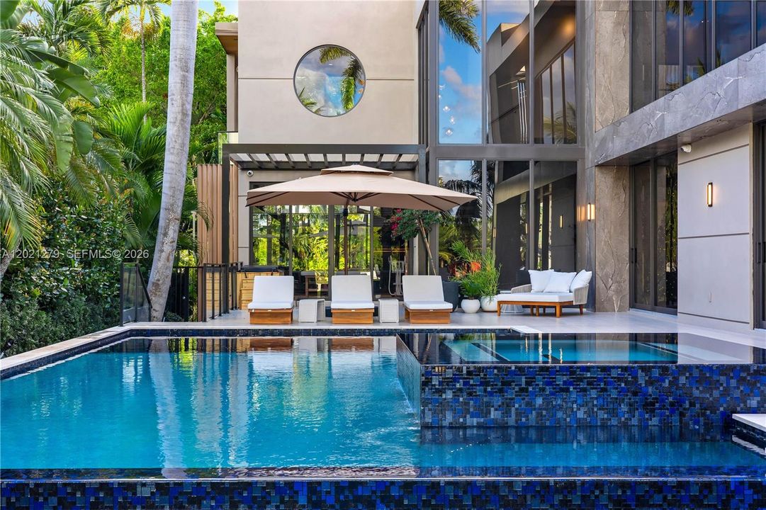 Oversized pool and elevated spa with blue mosaic tile waterfall spillover, teak chaise lounges, cantilever umbrella, woven lounge seating, large-format stone pool deck, floor-to-ceiling glass rear elevation, stone cladding, circular porthole window, and glass-enclosed wellness center beyond.