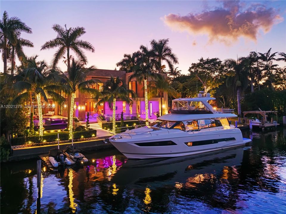 Ground-level dusk view from canal of private deep-water dock with large motor yacht, illuminated seawall, string-lit palms, manicured waterfront lawn, LED-accented outdoor living terrace, and full two-story lit rear elevation.