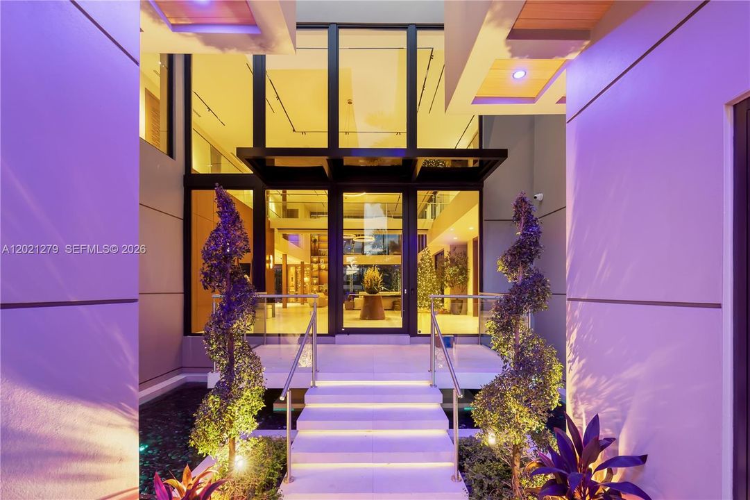 Double-height glass-and-steel entry with marble steps, stainless steel railings, spiral topiaries, and open view into grand two-story foyer interior.