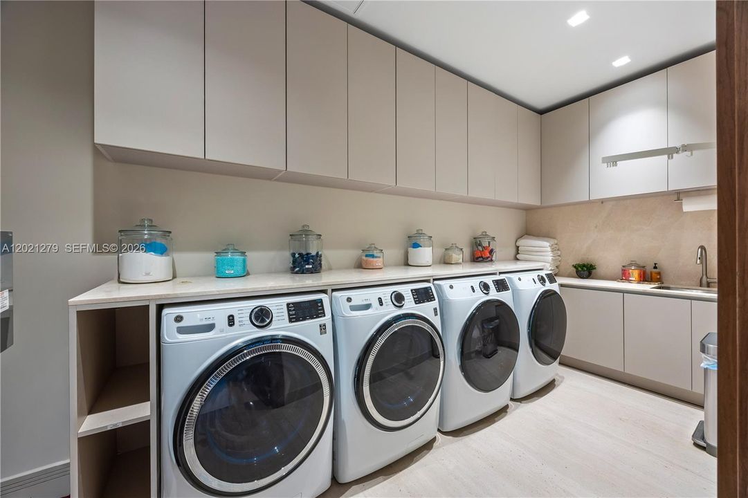 Large dedicated laundry room with dual washer and dryer sets, full-height flat-panel upper cabinetry, stone countertop, utility sink, light wood floors, and stone tile backsplash.