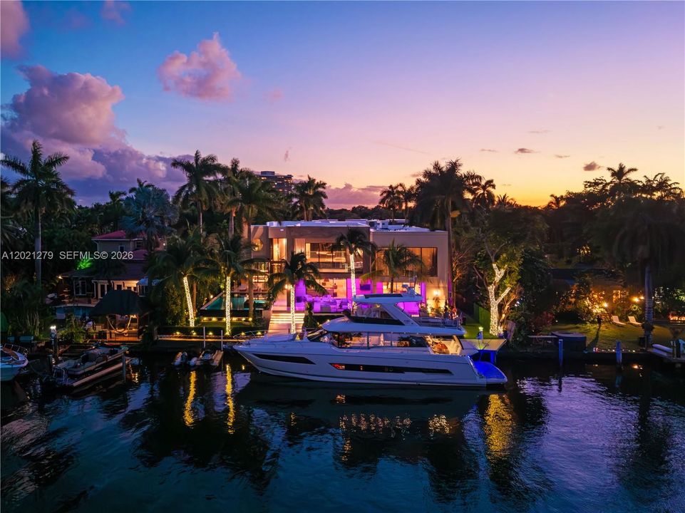 Aerial twilight view from canal showing two-story modern estate, private deep-water dock with large yacht, illuminated tropical landscaping, LED-lit palm trees, pool, covered outdoor living terrace, and dramatic purple and gold sunset sky.