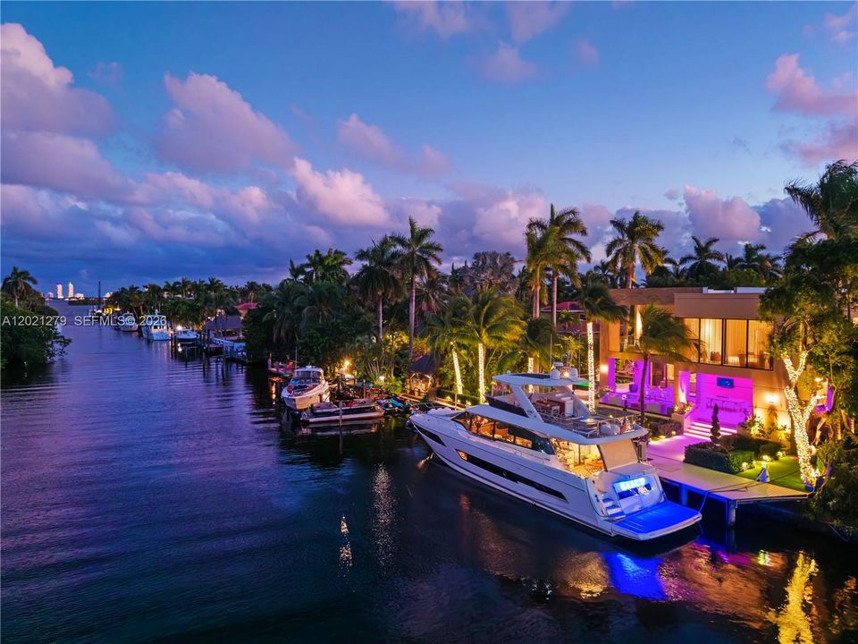 Aerial canal-level view at dusk featuring private deep-water dock, large motor yacht with underwater LED lighting, covered boat lift, illuminated waterfront terrace with purple LED accents, string-lit tropical palms, and canal stretching toward open bay.