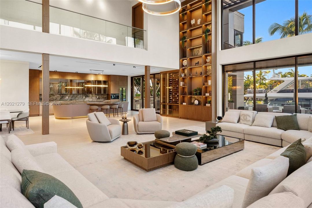 Wide-angle view of open-plan great room and chef's kitchen featuring curved fluted island with stone top, full-height illuminated walnut shelving, double-height glass walls with direct canal and yacht views, soaring ceilings, and light stone floors throughout.