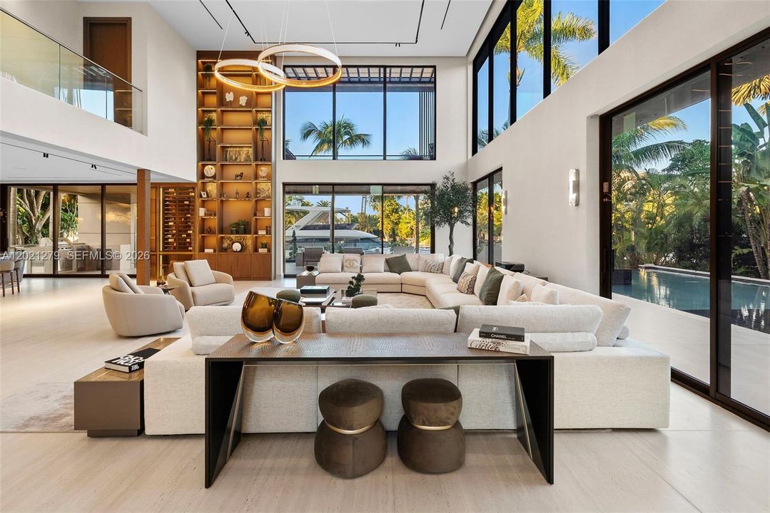 Sun-filled double-height great room with oversized curved sectional, dual ring chandelier, full-height illuminated walnut shelving, wine display, floor-to-ceiling glass with direct pool and waterfront views, and open second-floor balcony with glass railing.