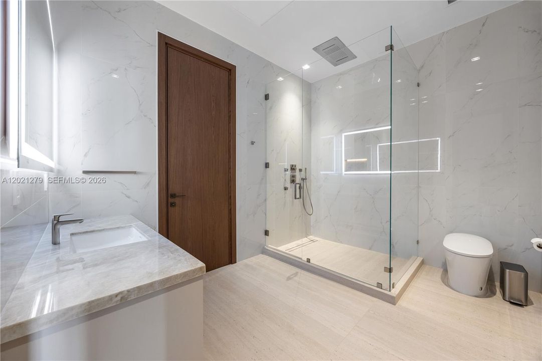 Modern guest bathroom with full-height white marble tile walls and floors, frameless glass walk-in shower, illuminated recessed niche, multi-function shower fixtures, undermount sink, stone vanity countertop, backlit mirror, and walnut door.