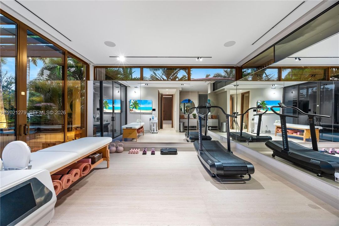 Full private gym and wellness suite with treadmills, kettlebells, yoga mats, massage table, full-wall mirror, wall-mounted TV, light wood floors, clerestory windows, floor-to-ceiling bronze-framed glass walls, and adjacent treatment or steam room.