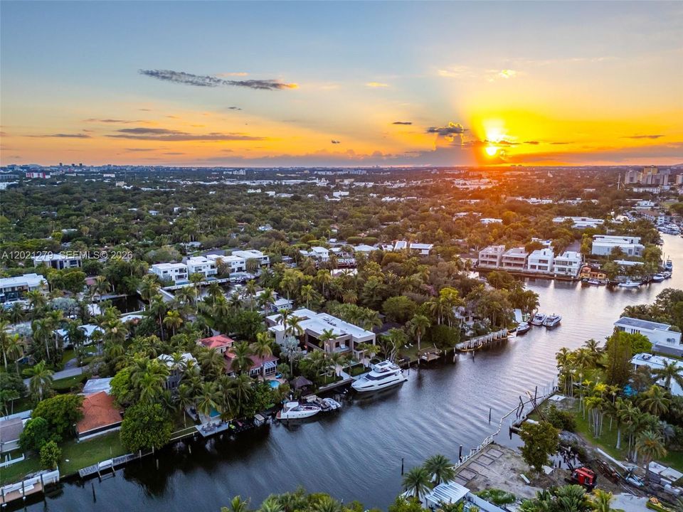 Sweeping aerial sunset view of deep-water canal with property and private dock visible, large motor yacht docked, lush tropical neighborhood, and vibrant golden sunset sky over the Miami skyline in the distance.