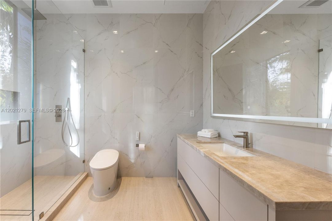 En-suite bath with full-height white marble tile walls and floors, frameless glass walk-in shower, smart toilet, floating vanity with warm stone countertop, undermount sink, brushed nickel fixtures, and backlit LED mirror.