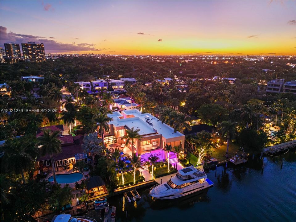 Aerial dusk view of modern waterfront estate with illuminated pool, covered loggia, manicured grounds, private dock with yacht, programmable LED lighting, and sweeping South Florida skyline and city lights backdrop.