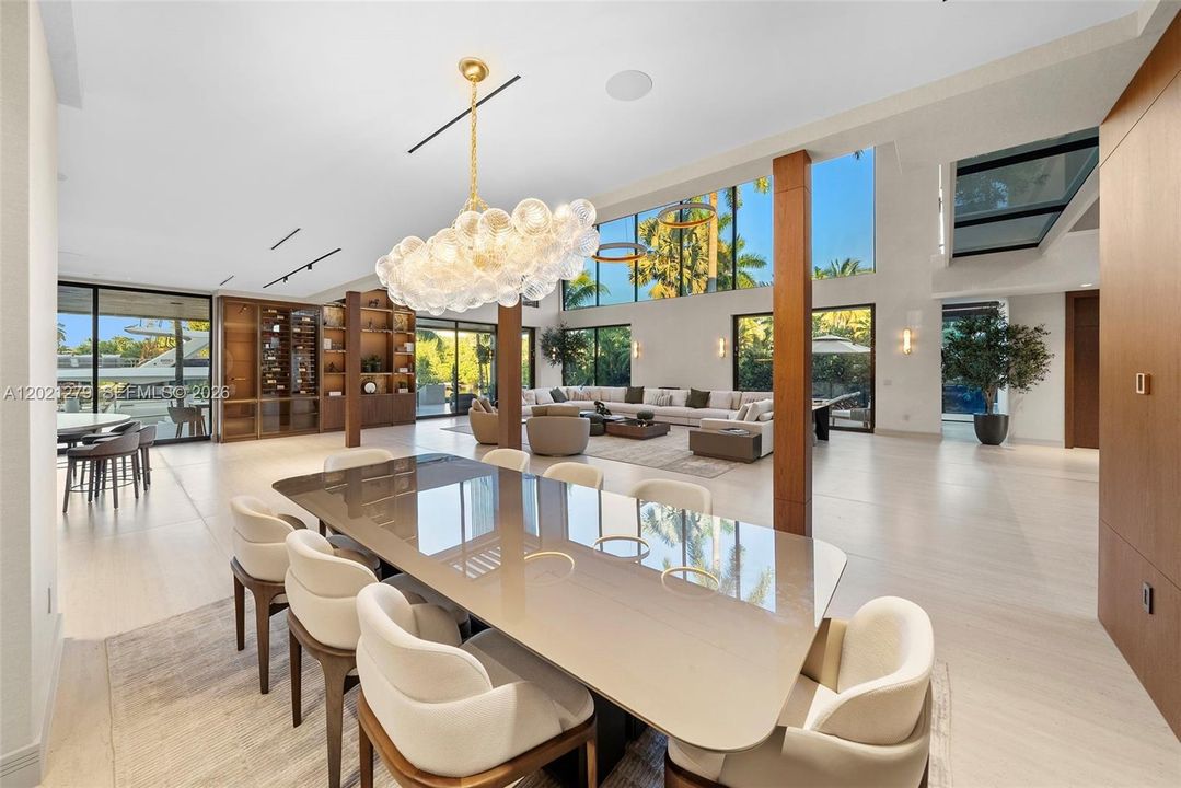 Open-plan formal dining area with large stone-top dining table, upholstered walnut chairs, oversized globe chandelier with brass stem, floor-to-ceiling glass with waterfront views, full-height walnut shelving with wine display, and adjacent great room.