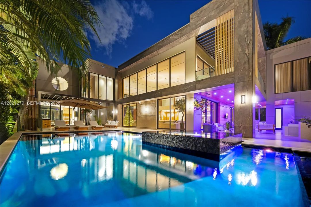 Night view of full rear elevation with illuminated pool, dark mosaic spa, stone-clad two-story facade, floor-to-ceiling glass, circular porthole window, second-level balcony with pergola, covered loggia, chaise lounges, and vibrant purple LED ambient lighting.