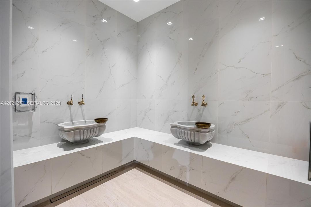 Spa-quality steam room with full-height polished white marble tile walls, corner L-shaped bench, dual ornate carved white vessel sinks, antique brass wall-mount faucets, linear floor drain, and digital climate control panel.