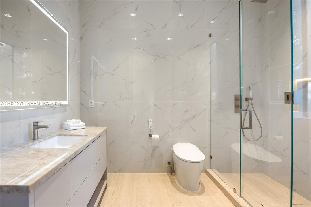 Clean modern en-suite bath featuring full-height white marble tile surround, frameless glass shower with hand-held fixture, smart toilet, floating white vanity, warm stone countertop, undermount sink, and backlit LED mirror.