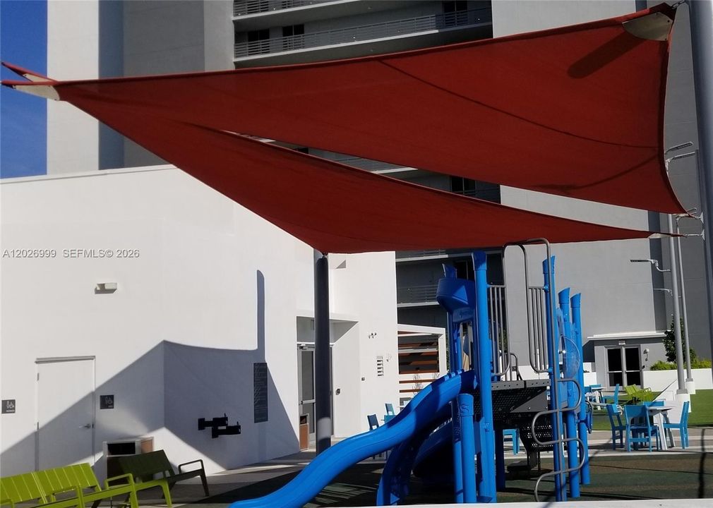 A shaded outdoor recreation area with modern play structures, including slides and climbing elements, set beneath red canopy sails. Surrounding seating and open space provide a comfortable area for recreation within the community grounds.