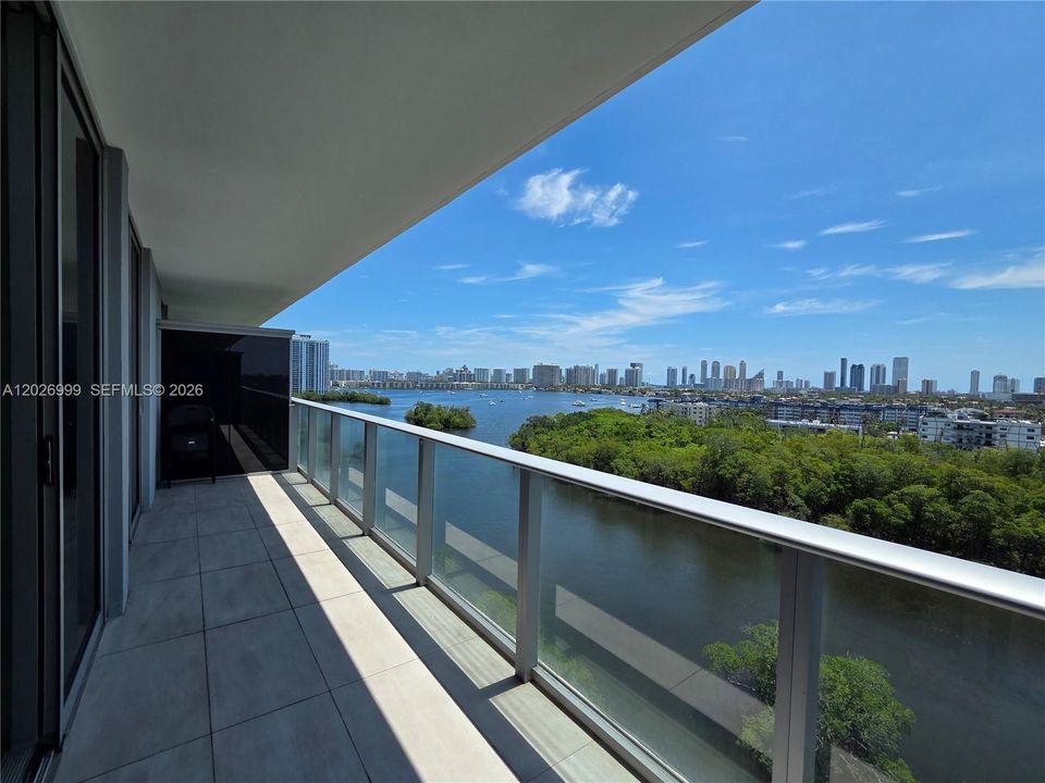 Spacious balcony with glass railings and sweeping views over the waterway, treetops, and skyline. The long outdoor area extends the living space with a scenic backdrop.