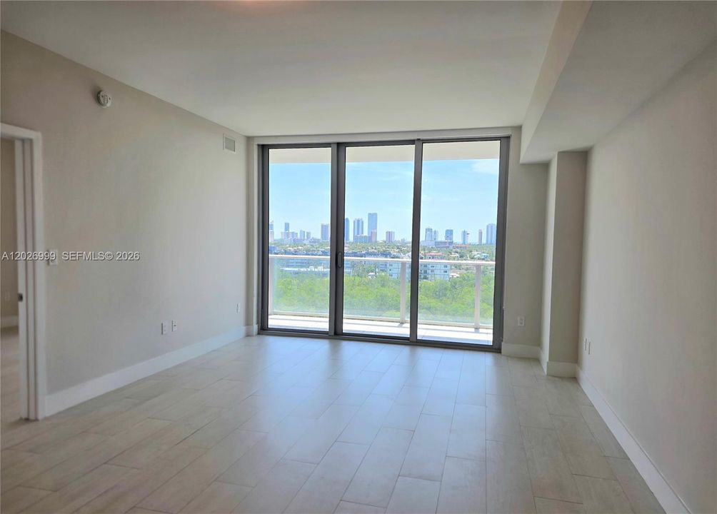 Bright open living area featuring large-format tile flooring and expansive sliding glass doors that open to a private balcony. Natural light fills the space while showcasing wide water and city skyline views.