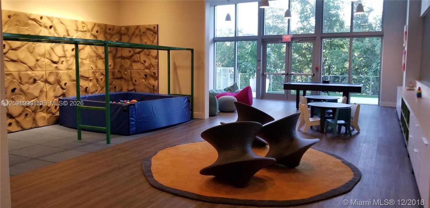 The children’s playroom features a climbing wall, activity tables, lounge seating, and floor-to-ceiling windows. This bright amenity space offers a dedicated area for play and recreation.