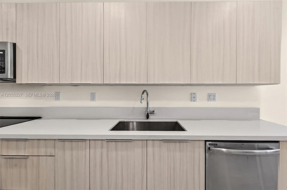 Sleek modern kitchen with light wood-grain cabinetry and crisp white quartz countertops. A deep stainless steel sink pairs with a pull-down faucet for added convenience, while stainless appliances complete the contemporary look.