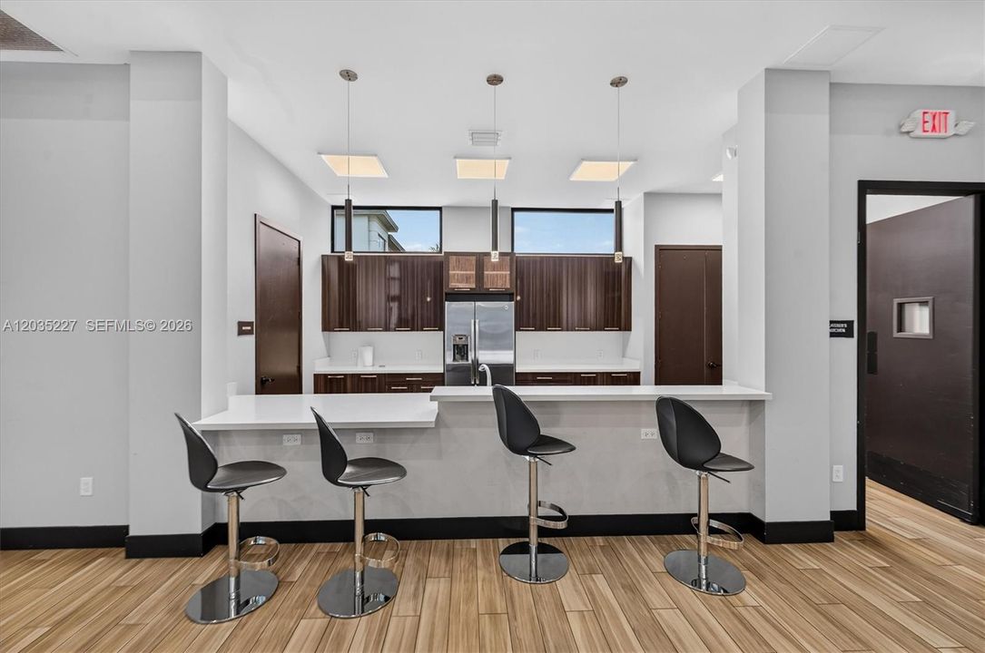Trendy community kitchen lounge featuring rich espresso cabinetry and a stylish bar with sleek black stools. Pendant lighting and clerestory windows add a chic, sophisticated touch above the warm wood flooring.