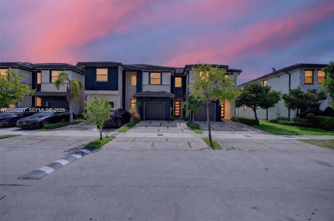 Twilight exterior shot — modern townhome glowing warmly against a vivid pink-and-blue sunset sky, navy and white facade, manicured landscaping, and clean curb appeal.