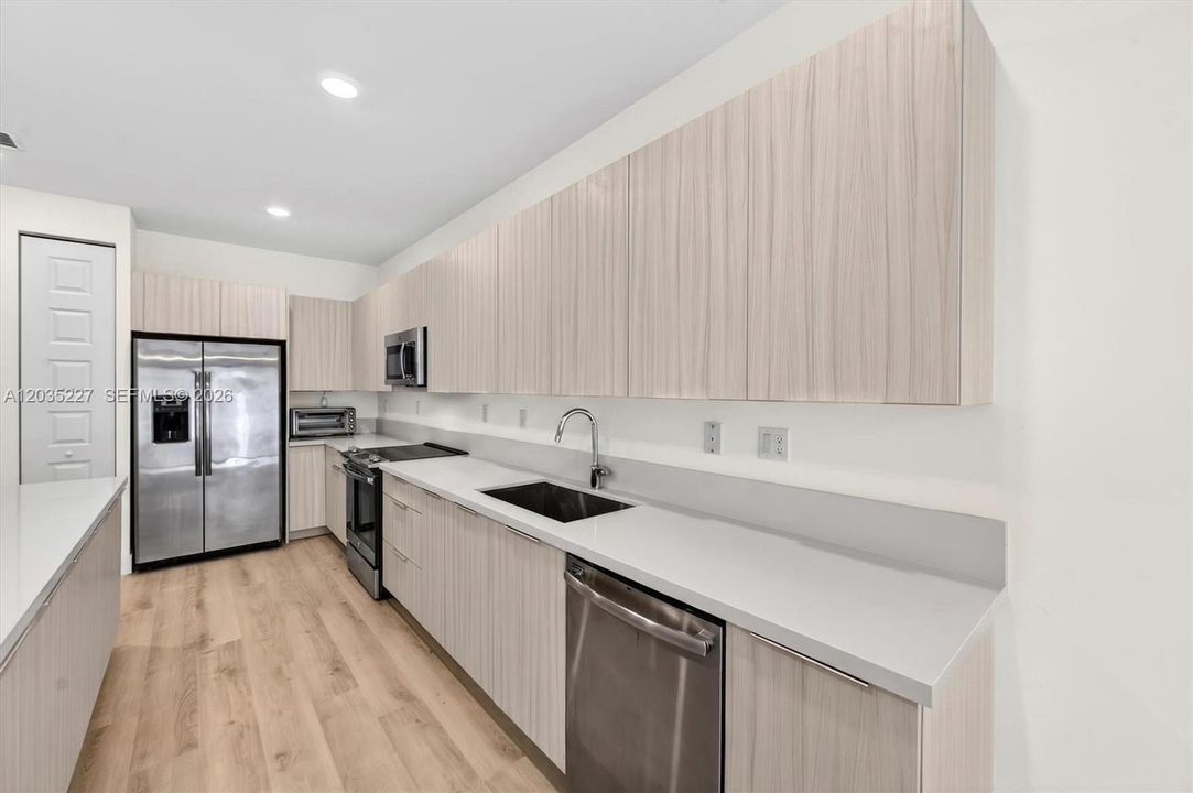 Galley-style kitchen with extensive counter space and floor-to-ceiling wood-grain cabinetry. Stainless steel appliances, including a side-by-side fridge and dishwasher, blend seamlessly with the sleek quartz countertops.
