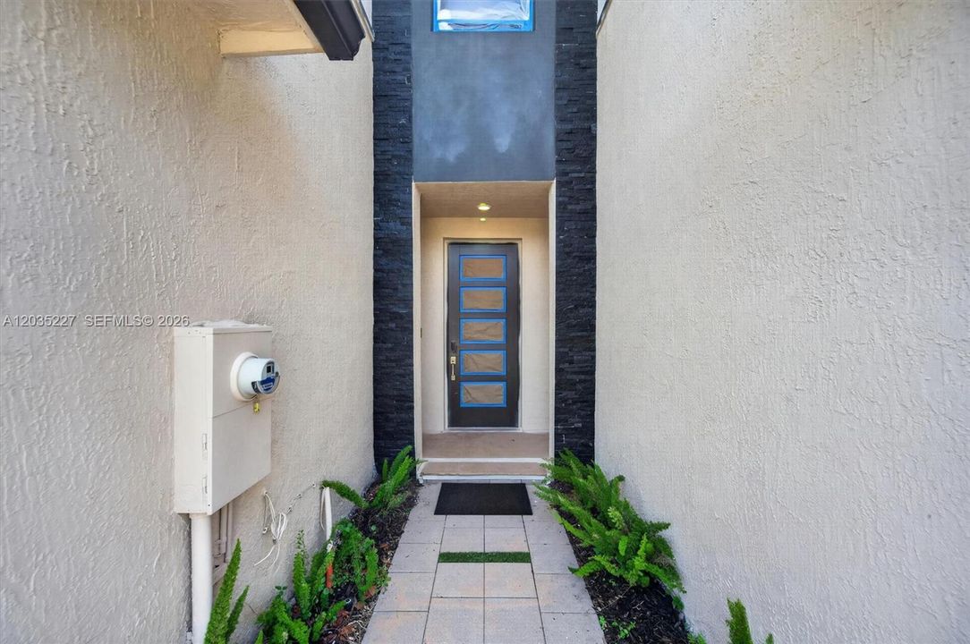 Private front entry walkway — framed by stacked stone and clean stucco, leading to a recessed door with soft accent lighting and lush green border plantings.