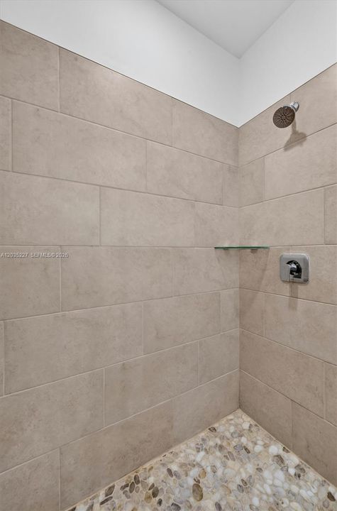 Spa-style walk-in shower finished with large-format tile and a natural pebble-stone floor for a luxurious, organic touch. A floating glass shelf and chrome fixtures complete the clean, modern design.