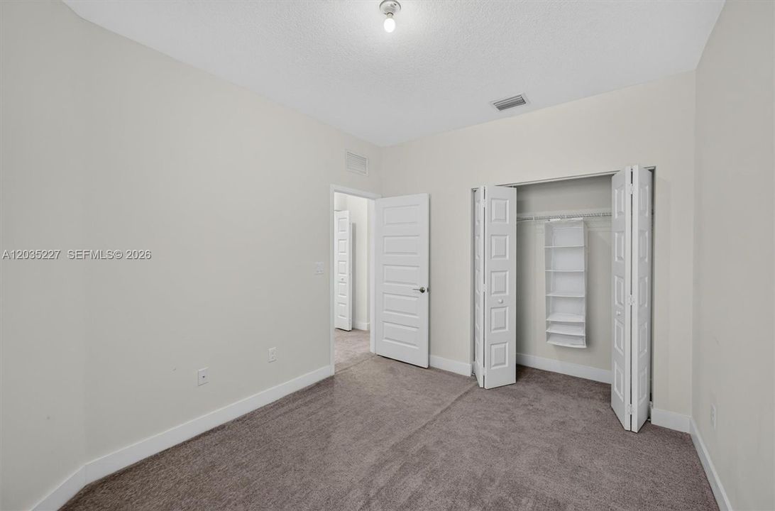 Bright secondary bedroom featuring soft carpeting and warm neutral walls. A spacious bi-fold closet with built-in shelving and hanging rods offers excellent storage. Easy access to the hallway makes this room both functional and inviting.