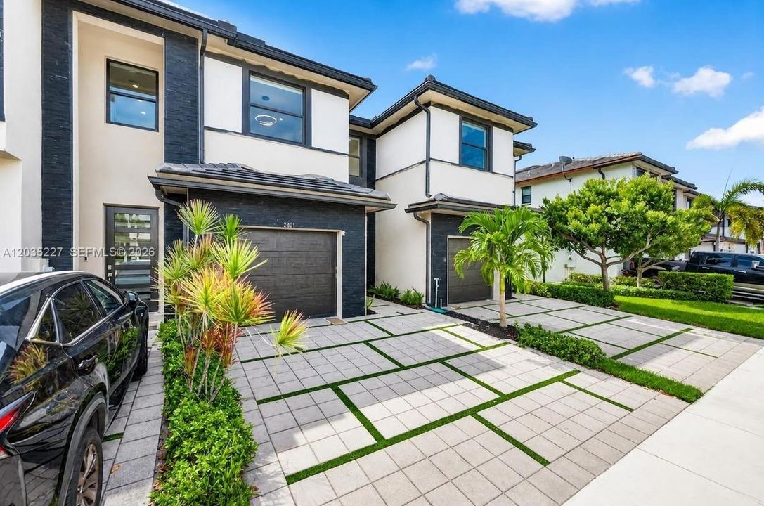 Front exterior view — contemporary two-story townhome with charcoal stone accents, white stucco, dark-frame impact windows, and a striking geometric paver driveway lined with palms and tropical landscaping.