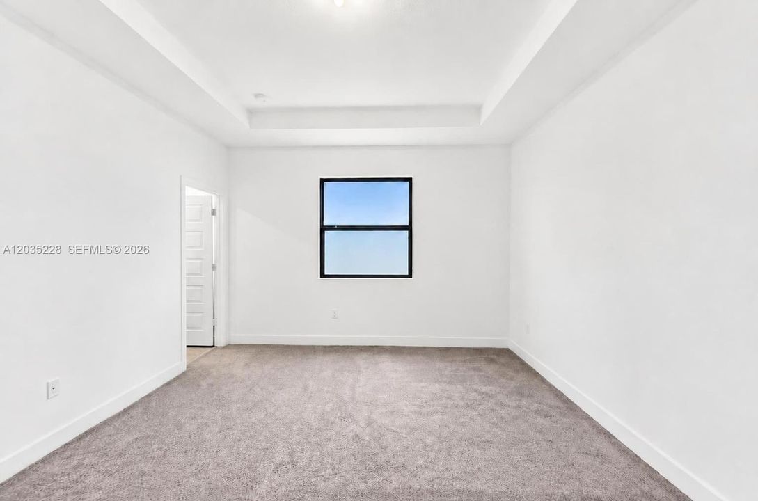Spacious bedroom with plush carpeting and a tray ceiling that adds architectural detail. A large window brings in natural light, complemented by a cordless shade for privacy. Neutral tones throughout offer a blank canvas for personal style.