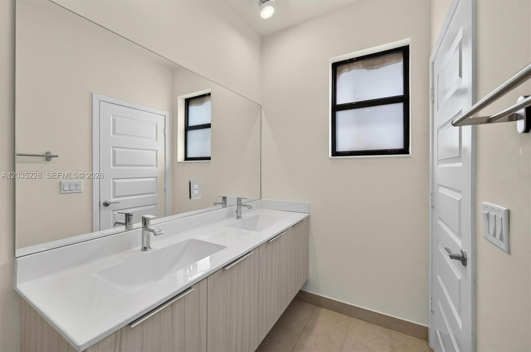 Spa-inspired bathroom featuring a long double-sink vanity with quartz countertops and a frameless mirror that doubles the natural light. Wood-grain cabinetry and tile flooring add warmth to this functional, well-appointed space.
