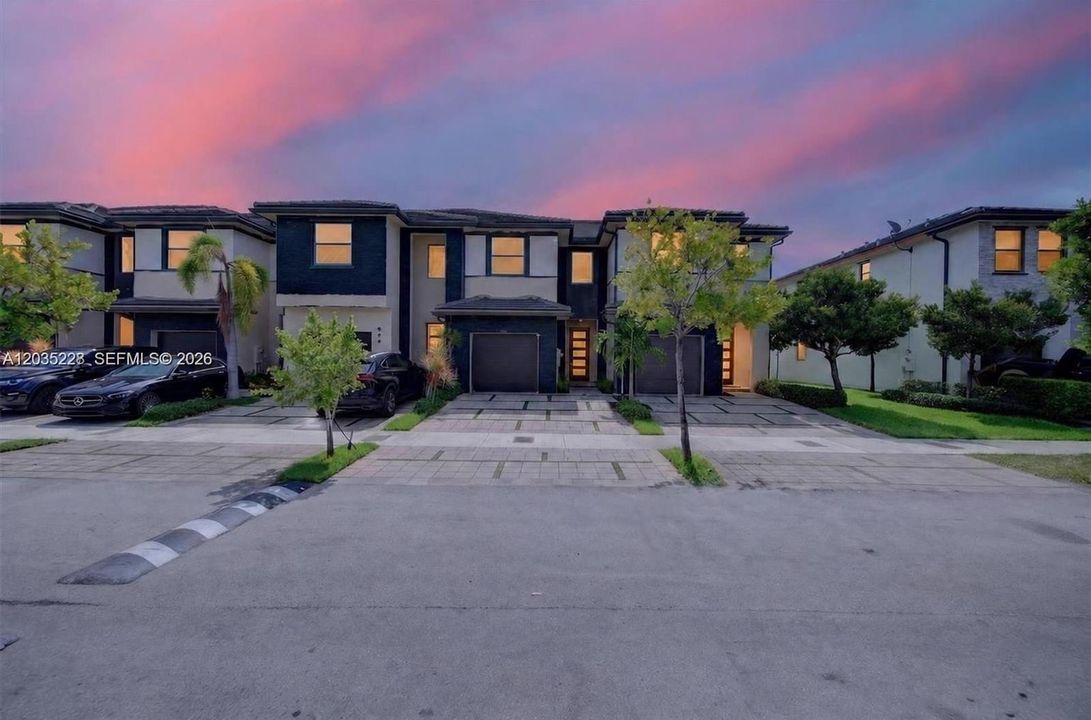 Twilight exterior shot — modern townhome glowing warmly against a vivid pink-and-blue sunset sky, navy and white facade, manicured landscaping, and clean curb appeal.