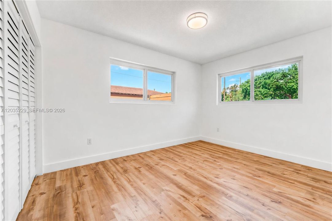 Sun-filled bedroom with warm wood-look flooring, dual windows overlooking lush tropical greenery, and a plantation shutter closet.