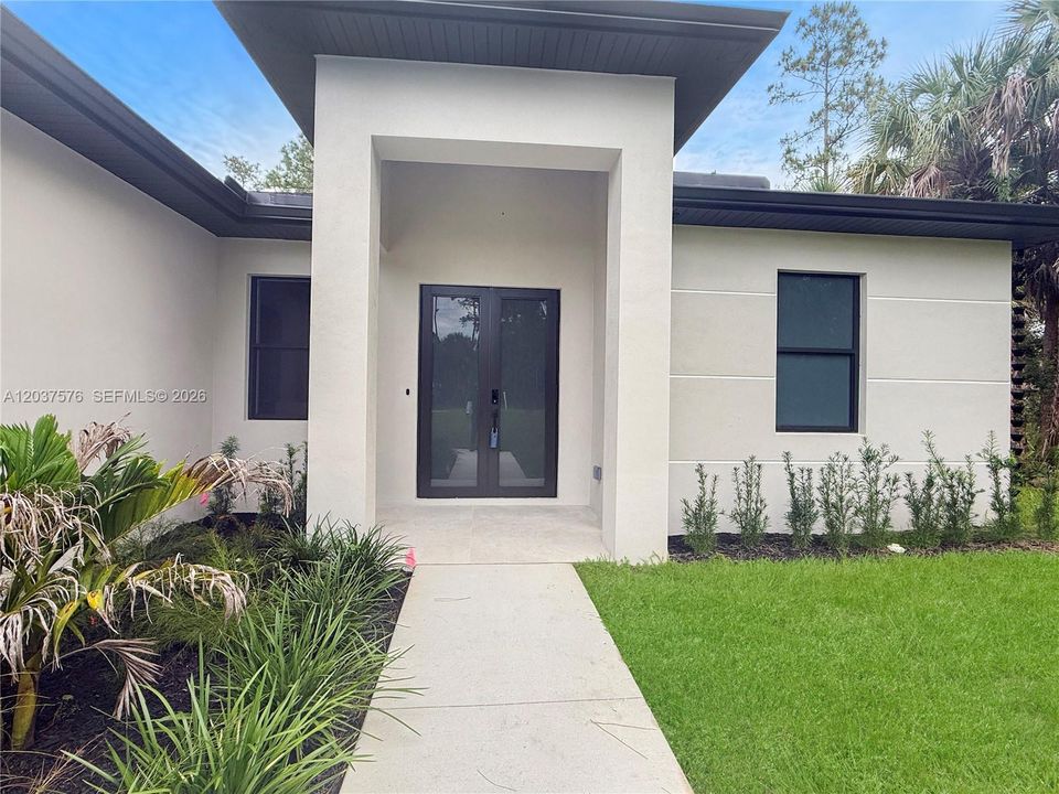Modern stucco exterior featuring a covered entry, dark-framed double entry doors, dark-framed windows, a concrete walkway, and landscaped garden beds