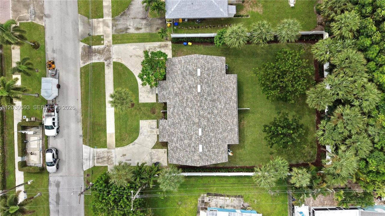 Aerial view featuring a paved circular driveway, a shingle-style roof, and a fenced-in lawn with mature landscaping