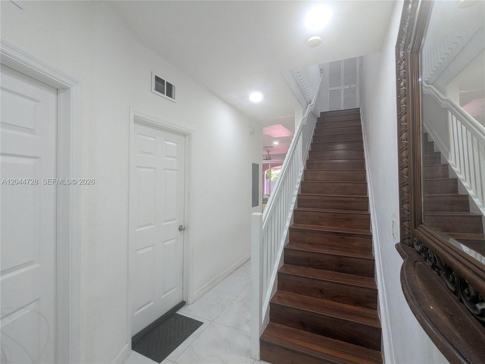 Bright interior hallway with white tile flooring and dark staircase. Clean layout with multiple doors and a large decorative mirror enhancing the space.