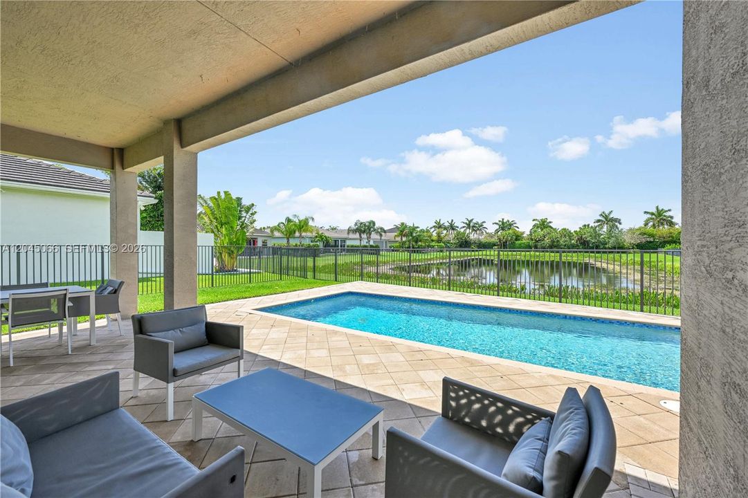Covered patio with stone-paver flooring, rectangular swimming pool, and metal perimeter fencing overlooking a pond