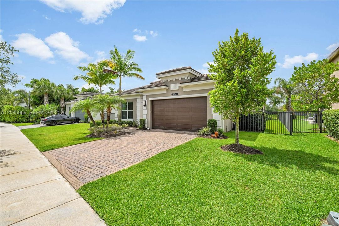 Single-story exterior featuring a paver driveway, attached two-car garage, manicured lawn, metal perimeter fencing, and established palm trees
