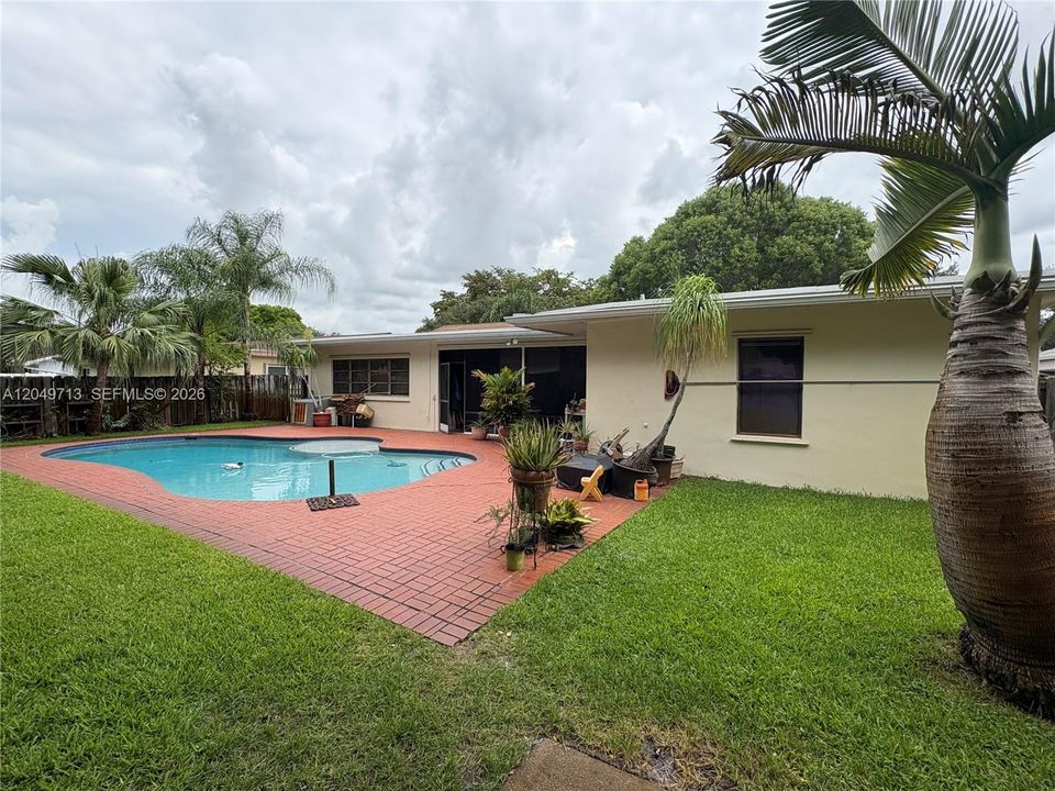 Backyard area featuring an inground swimming pool, red brick paver decking, manicured lawn, and mature palm trees