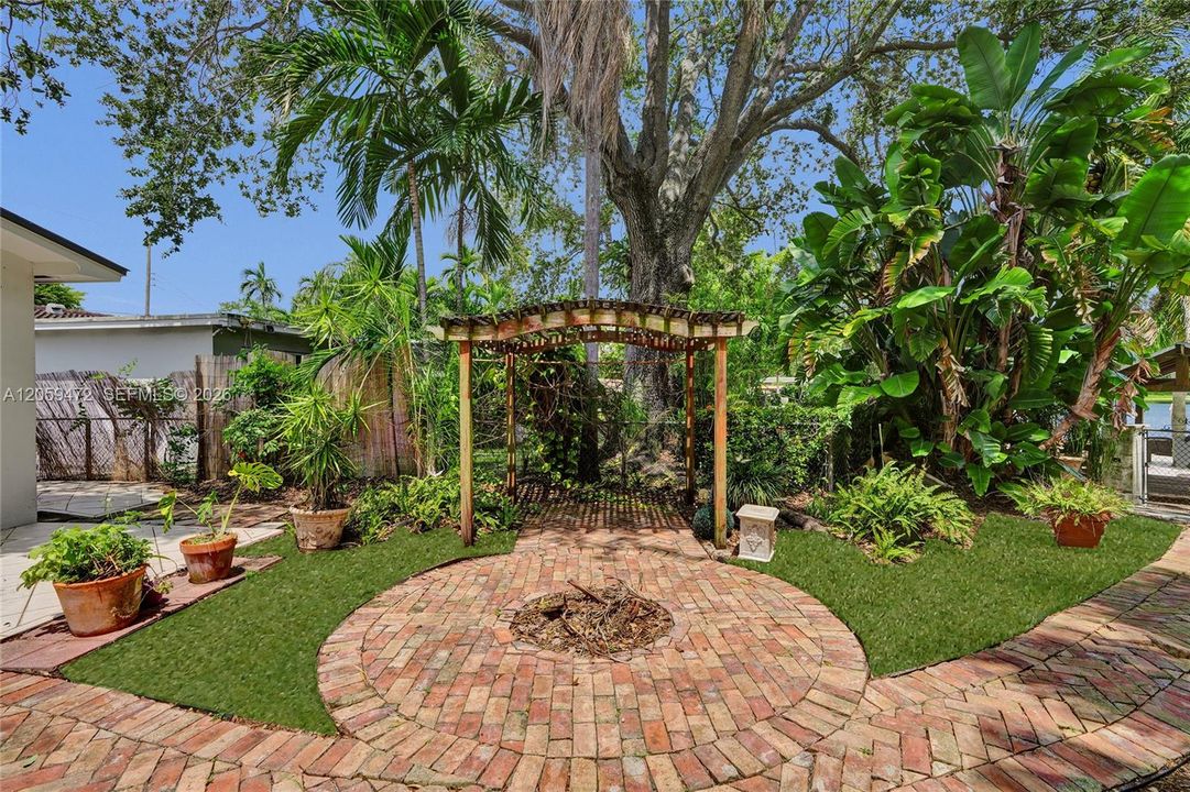 Lush, private lakefront backyard oasis — Charming brick-paved patio with circular fire pit area, framed by a rustic wood pergola and mature tropical landscaping including palms, banana trees, and ferns. Manicured lawn sections and potted greenery add color throughout, while a canopy of oak and palm trees provides natural shade. A gated path leads toward the water's edge, offering a serene, tropical retreat just steps from the lake.