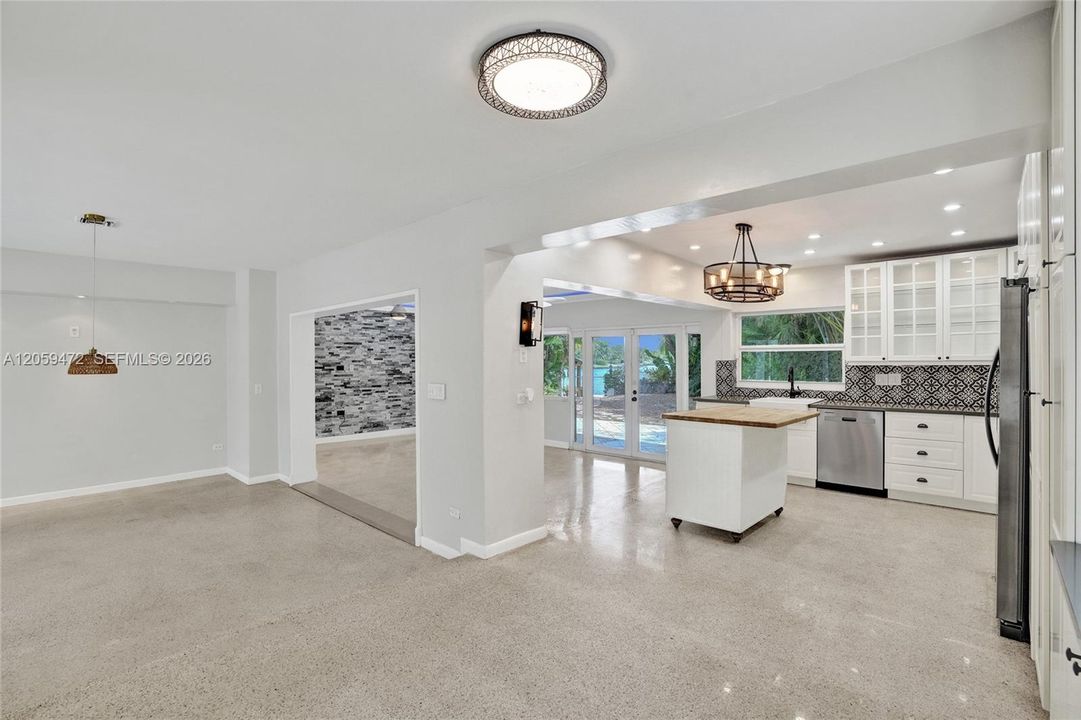 Wide view showing the seamless flow from the open living area into the kitchen, with a glimpse of the stacked stone accent wall beyond and French doors leading out to the lake. The kitchen island, glass-front cabinetry, and layered lighting (recessed, pendant, and drum chandelier) are all visible, highlighting the home's connected, entertainment-friendly layout.