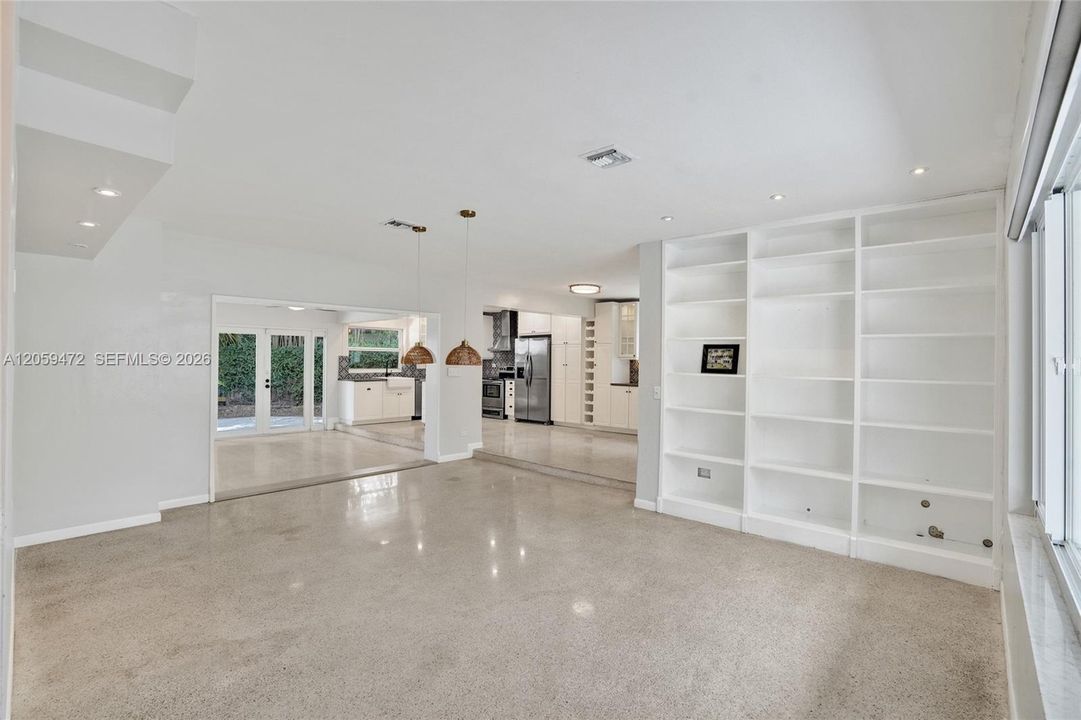 Expansive open-concept living area —boasts built-in bookshelves. Polished terrazzo flooring flows throughout this bright, multi-purpose space featuring custom built-in shelving, woven pendant lighting, and clear sightlines into the updated kitchen and adjacent family room. Impact windows and doors lead out to the backyard, filling the space with natural light. A flexible layout perfect for everyday living and entertaining alike.