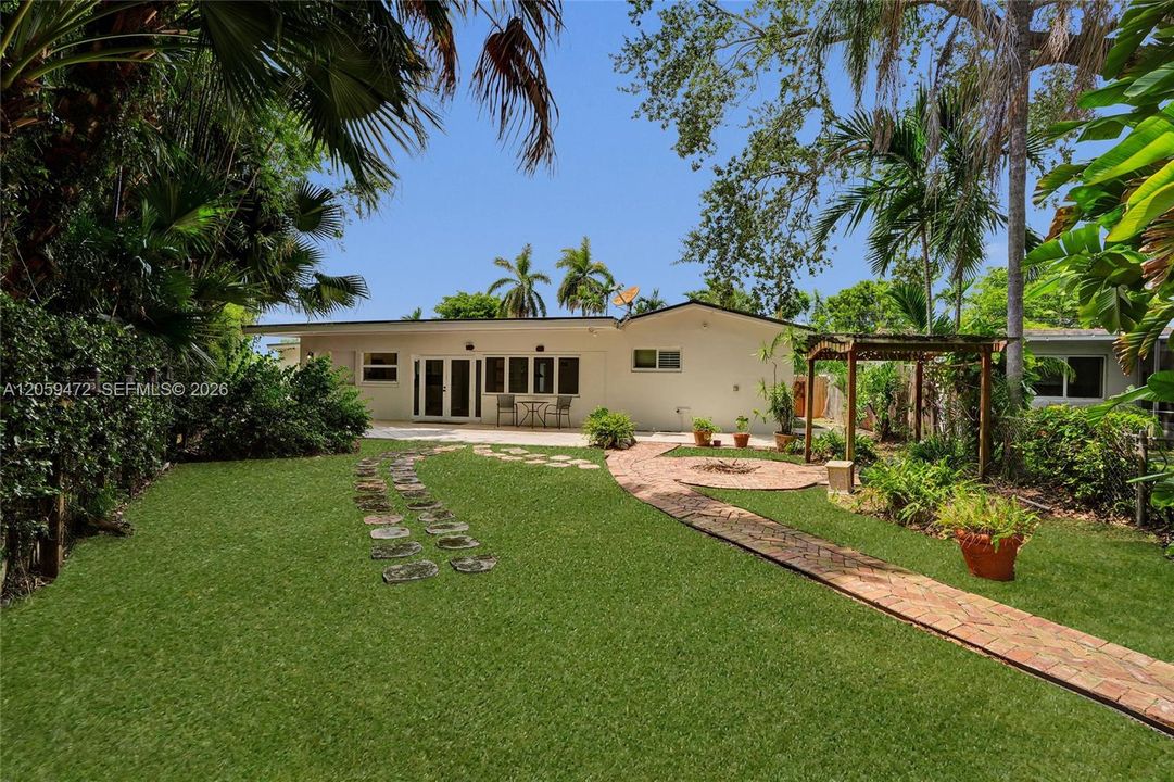 Sprawling, lush backyard framed by mature palms and tropical greenery, with a manicured lawn split by a whimsical stepping-stone path and a curved herringbone brick walkway leading to the covered patio. The single-story home features French doors and a private outdoor seating area, with a wood pergola and fire pit tucked to the side — a peaceful, tropical retreat with total privacy.