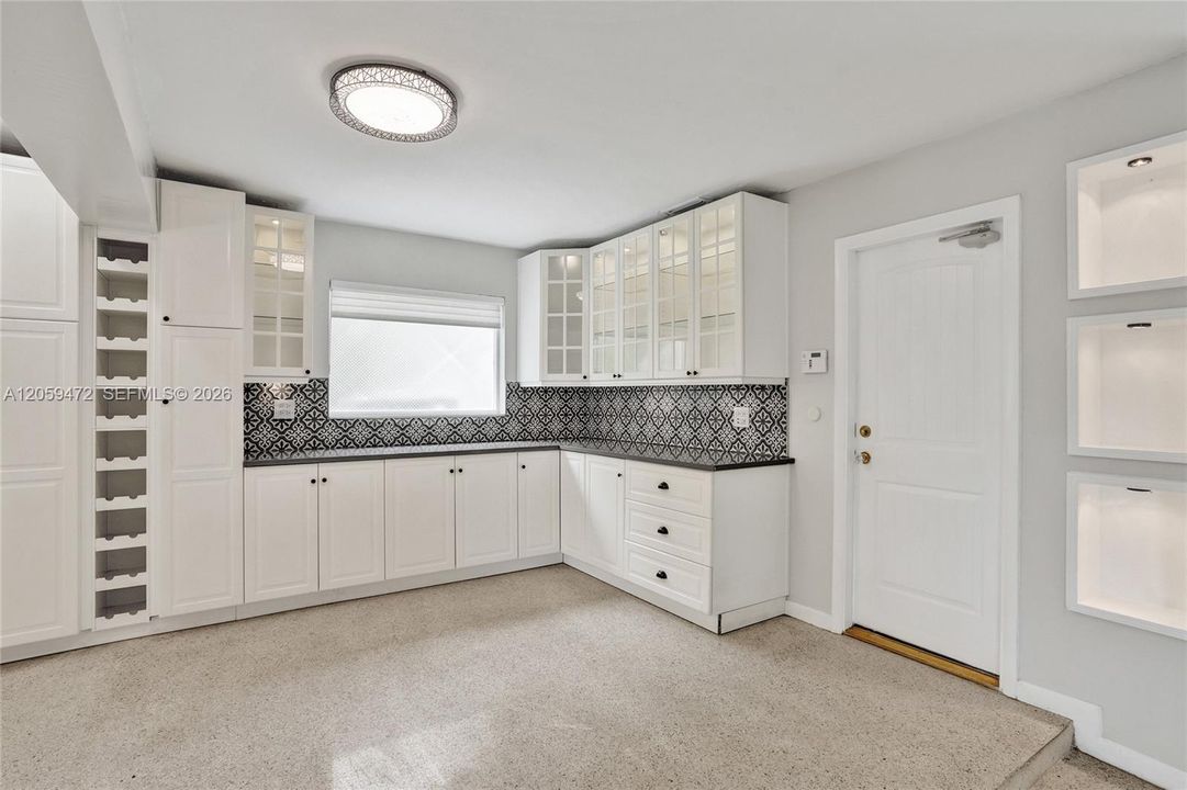 Butler's pantry / mudroom area just off the entry, featuring a wall of white cabinetry with glass-front uppers, a built-in wine rack, and matching black-and-white patterned tile backsplash. A modern woven flush-mount fixture lights the space, which connects directly to the 3-car garage — great extra storage adjacent to the kitchen.