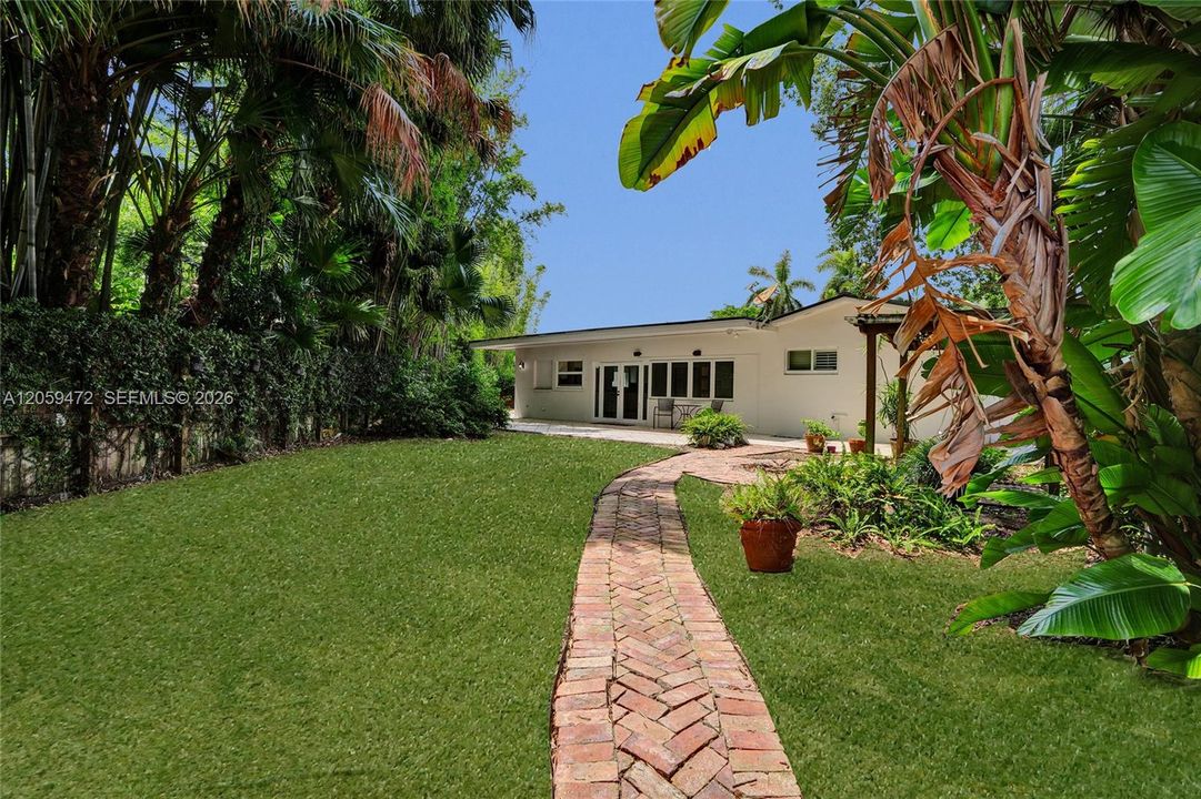 A closer view down the herringbone brick path, framed by banana trees and towering bamboo, leading back to the home's covered patio and French doors. Dense, layered tropical landscaping along the fence line creates a secluded, jungle-like feel while keeping the home and outdoor living space in clear view.