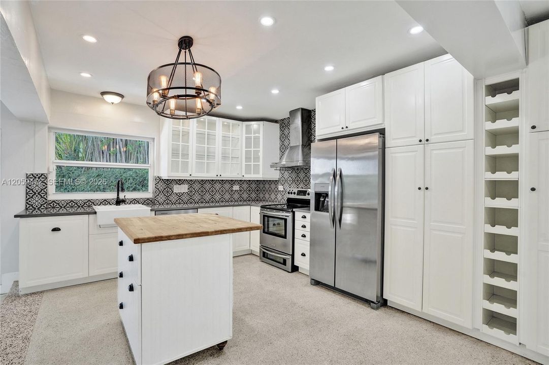 Full kitchen view showcasing the stainless French-door refrigerator, gas range with vent hood, farmhouse sink, and built-in wine storage — all framed by the bold patterned backsplash and warm Edison-bulb chandelier over the butcher-block island.